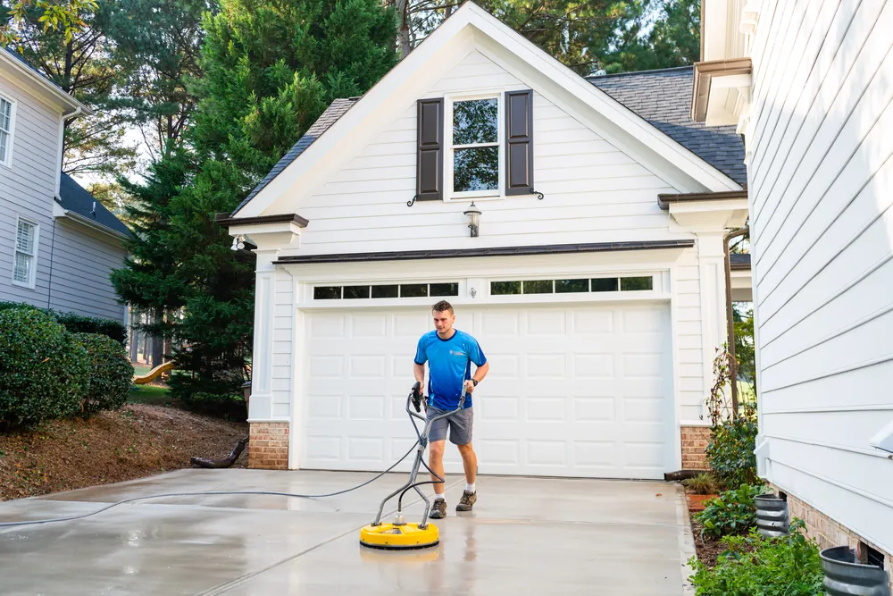 Technician running a surface cleaner across a concrete driveway in front of a white garage